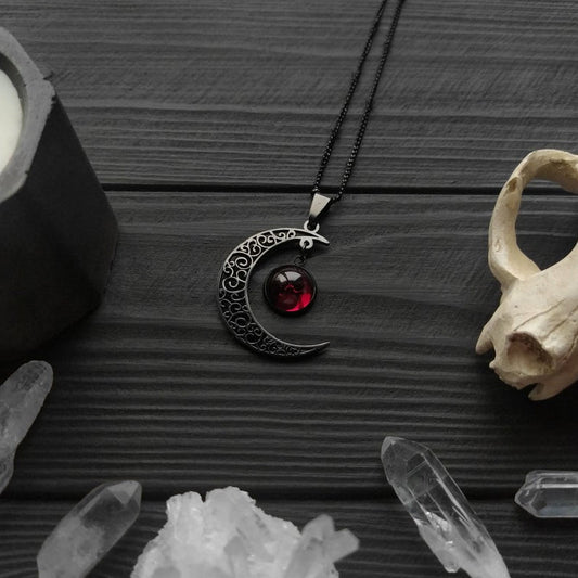 Black crescent moon necklace with red gemstone on a dark wooden surface with crystals and a candle.